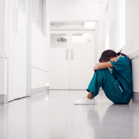 Stressed And Overworked Female Doctor Wearing Scrubs Sitting On Floor In Hospital Corridor